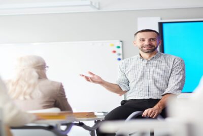 A man is speaking in front of a class while leaning on a desk.