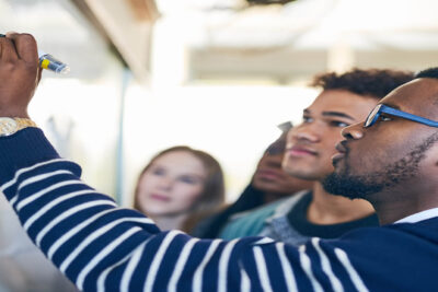Young man writing on a whiteboard while other students look on.